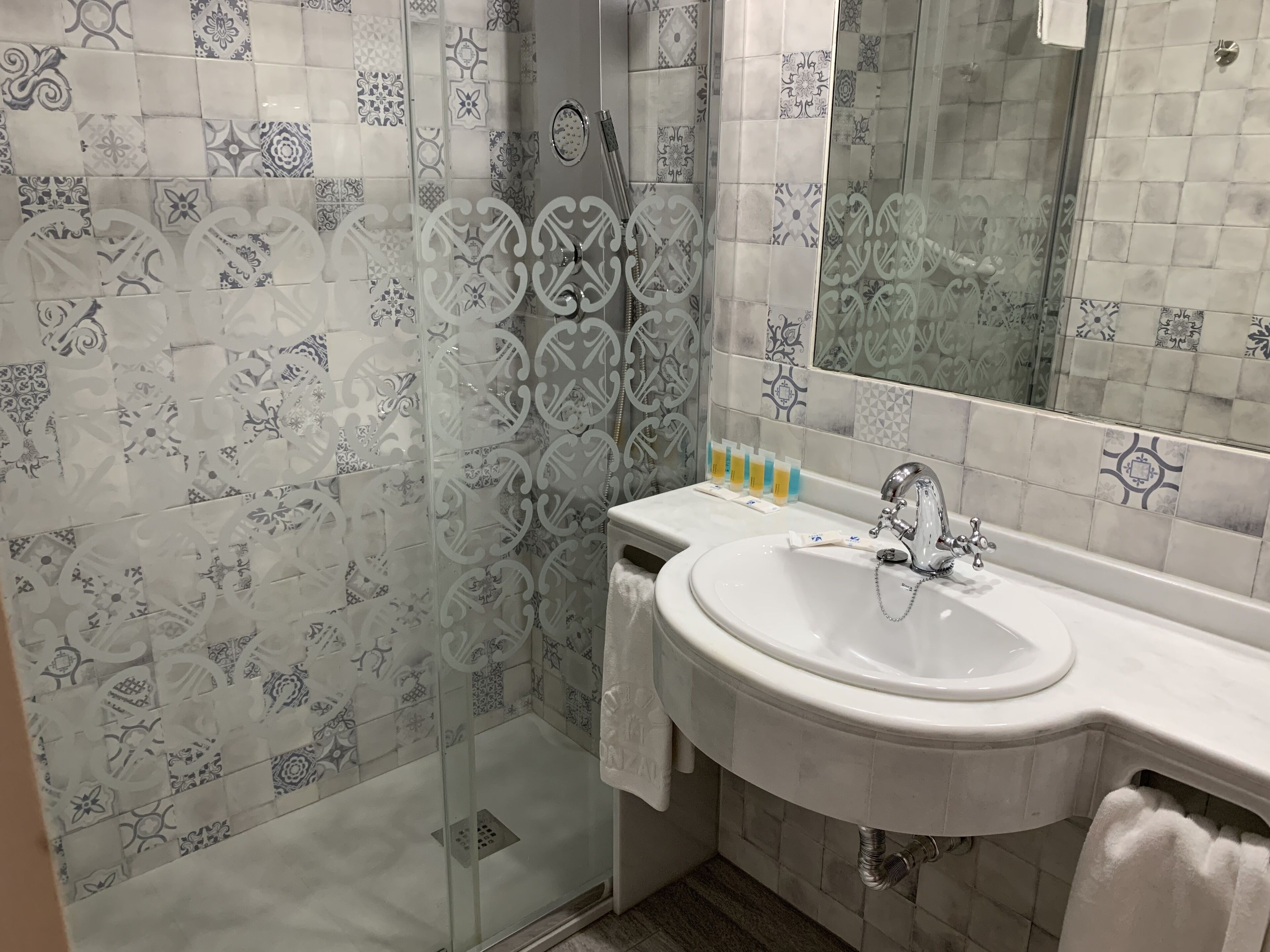 A bathroom with patterned tiles, a glass shower enclosure, a sink with toiletries, and a mirror.