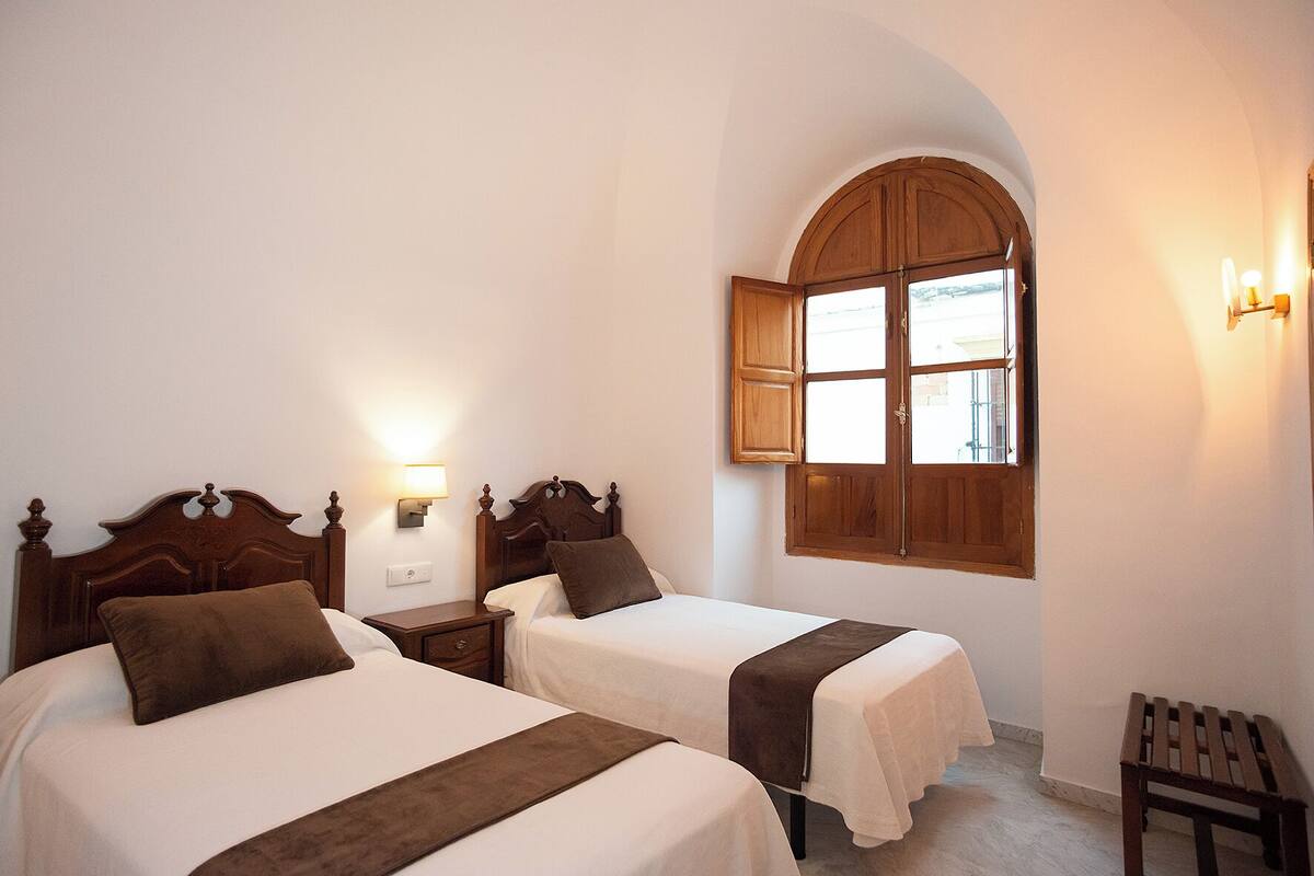 A hotel room with two beds, a wooden arched window, and a nightstand with a lamp.