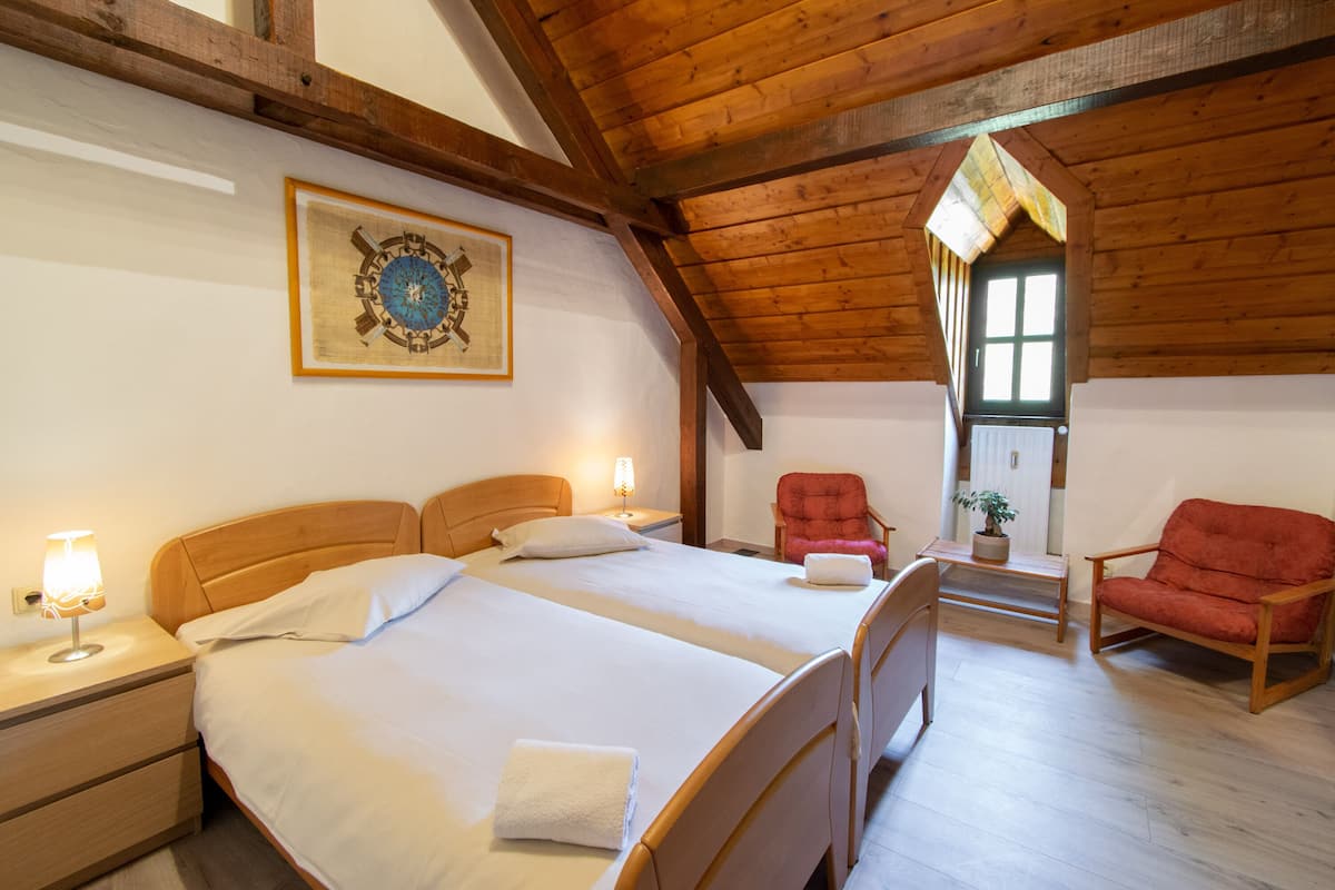 Ein Zimmer mit zwei Betten, Holz-Kopfteilen, einem roten Sessel, einem kleinen Tisch mit Pflanze und einem gerahmten Bild an der Wand.