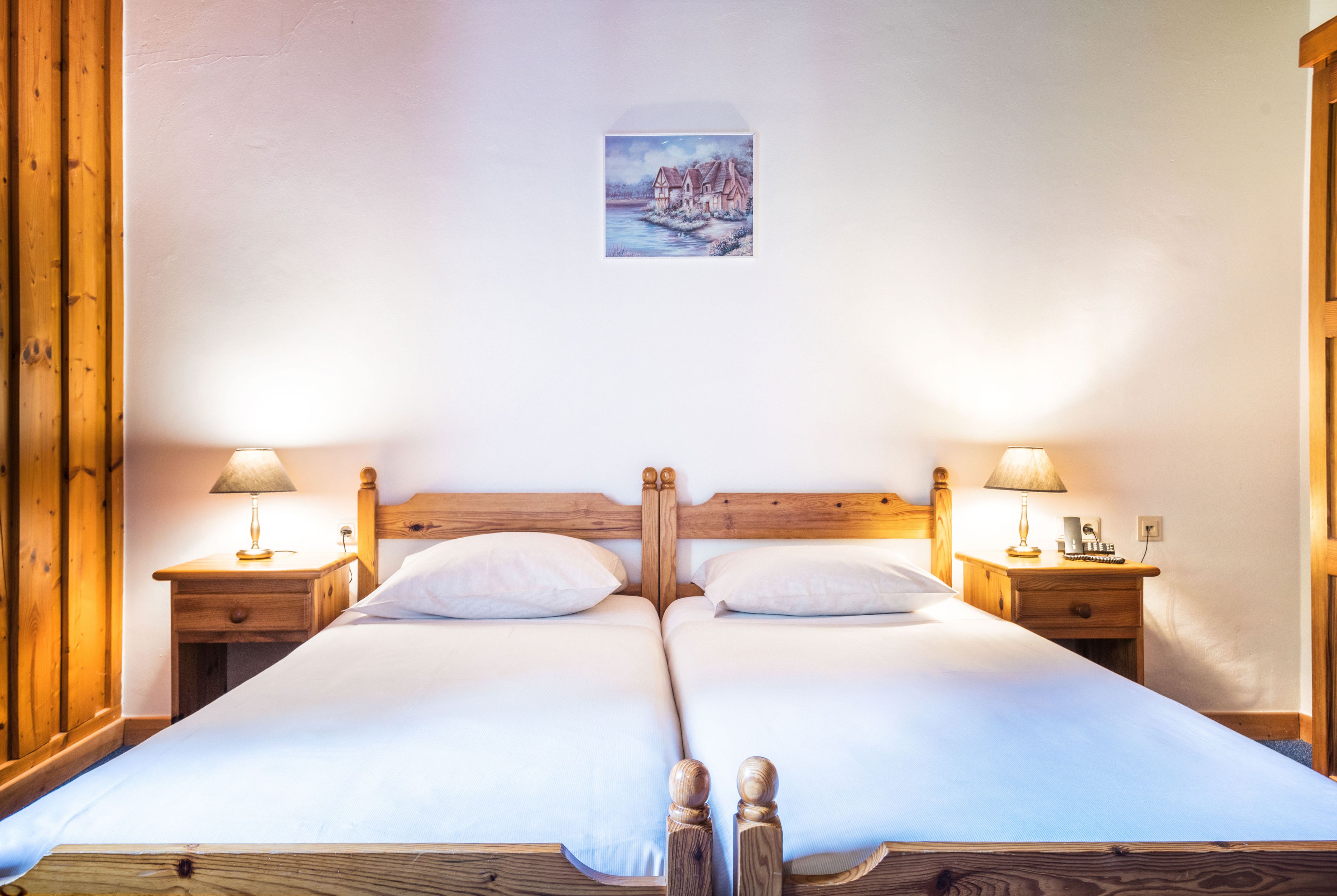 Uma cama de casal com cabeceira e peseira de madeira, duas mesas de cabeceira com abajures e um pequeno quadro na parede.