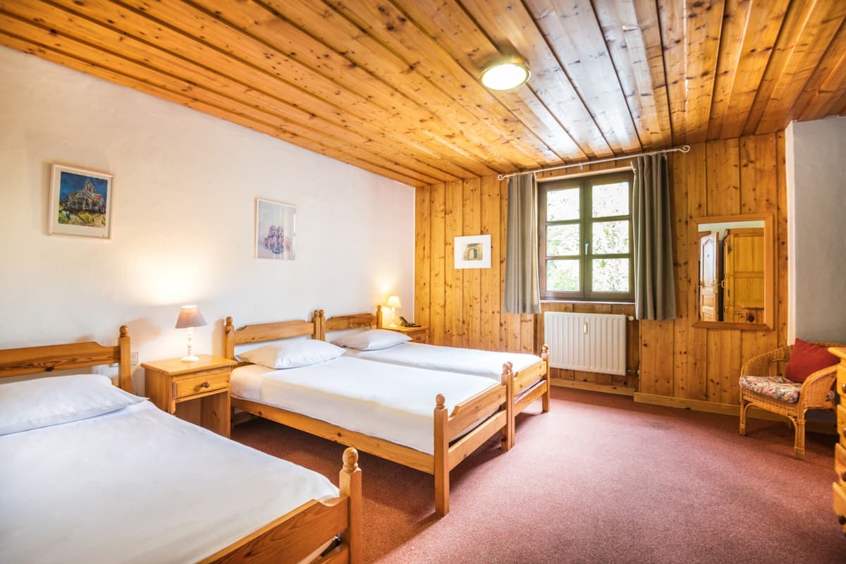 Eine Holzhütte mit drei Einzelbetten, einer Holzbalkendecke und einem Fenster mit Vorhängen.