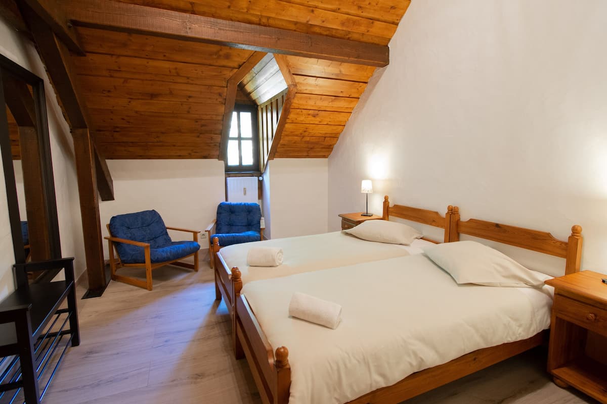 Ein Zimmer mit zwei hölzernen Betten, einem blauen Stuhl, einem Nachttisch aus Holz und einem Fenster mit Tageslicht.