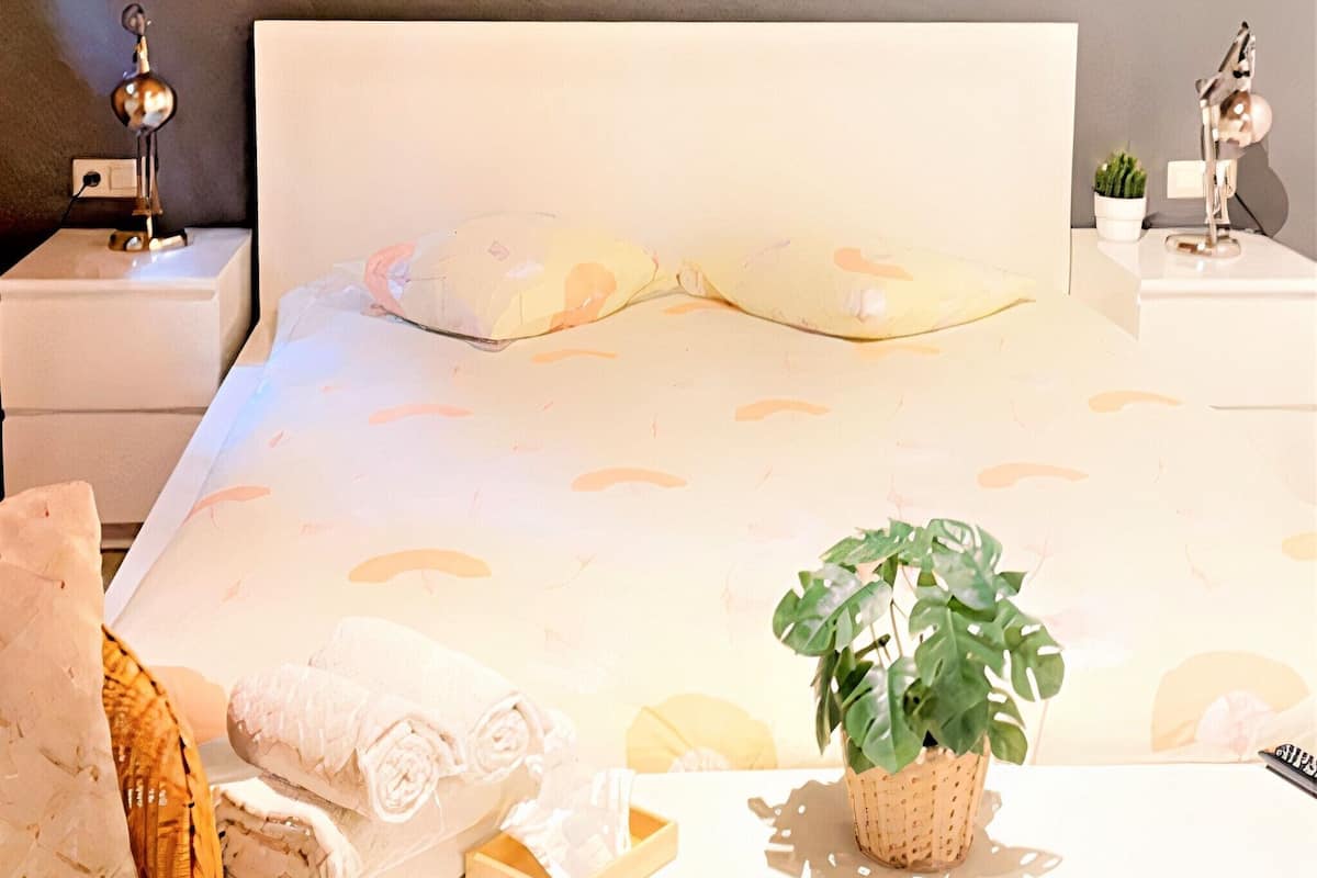 Ein ordentlich bezogenes Bett mit weißem Kopfteil, ein Nachttisch mit Lampe, ein kleiner Topfpflanze und eine Wandleuchte.