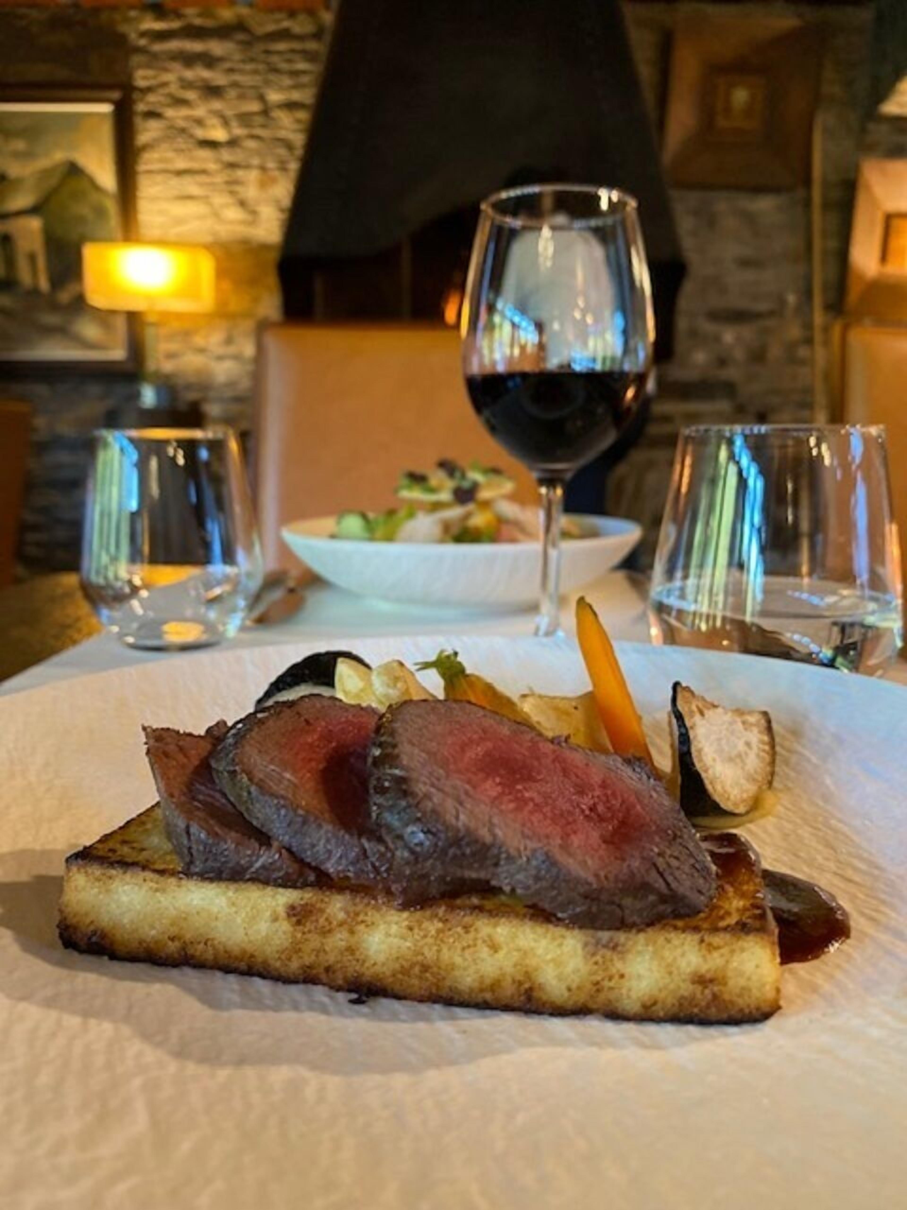 Un steak sur une tranche de pain grillé, accompagné d’un assortiment de légumes et d’un verre de vin rouge, le tout sur une table.