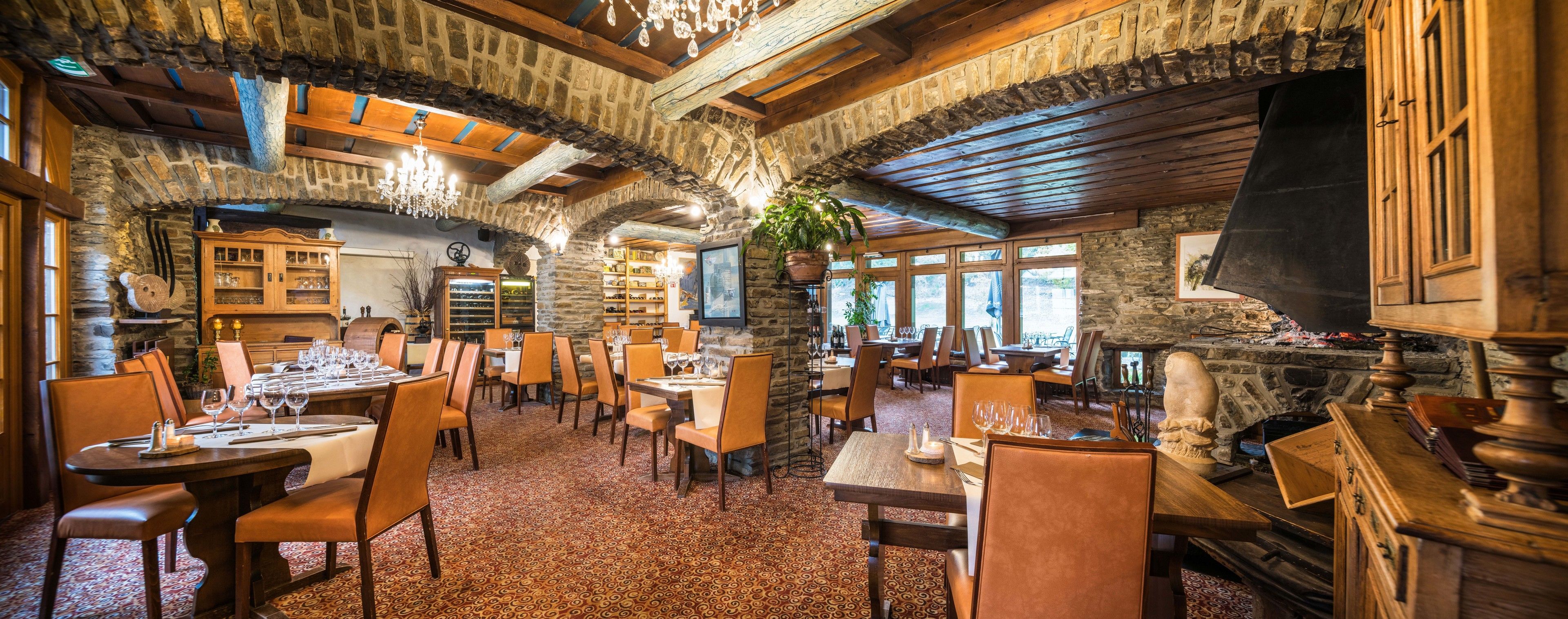 A rustic interior with stone walls, wooden beams, and a chandelier. Tables are set with wine glasses and plates.