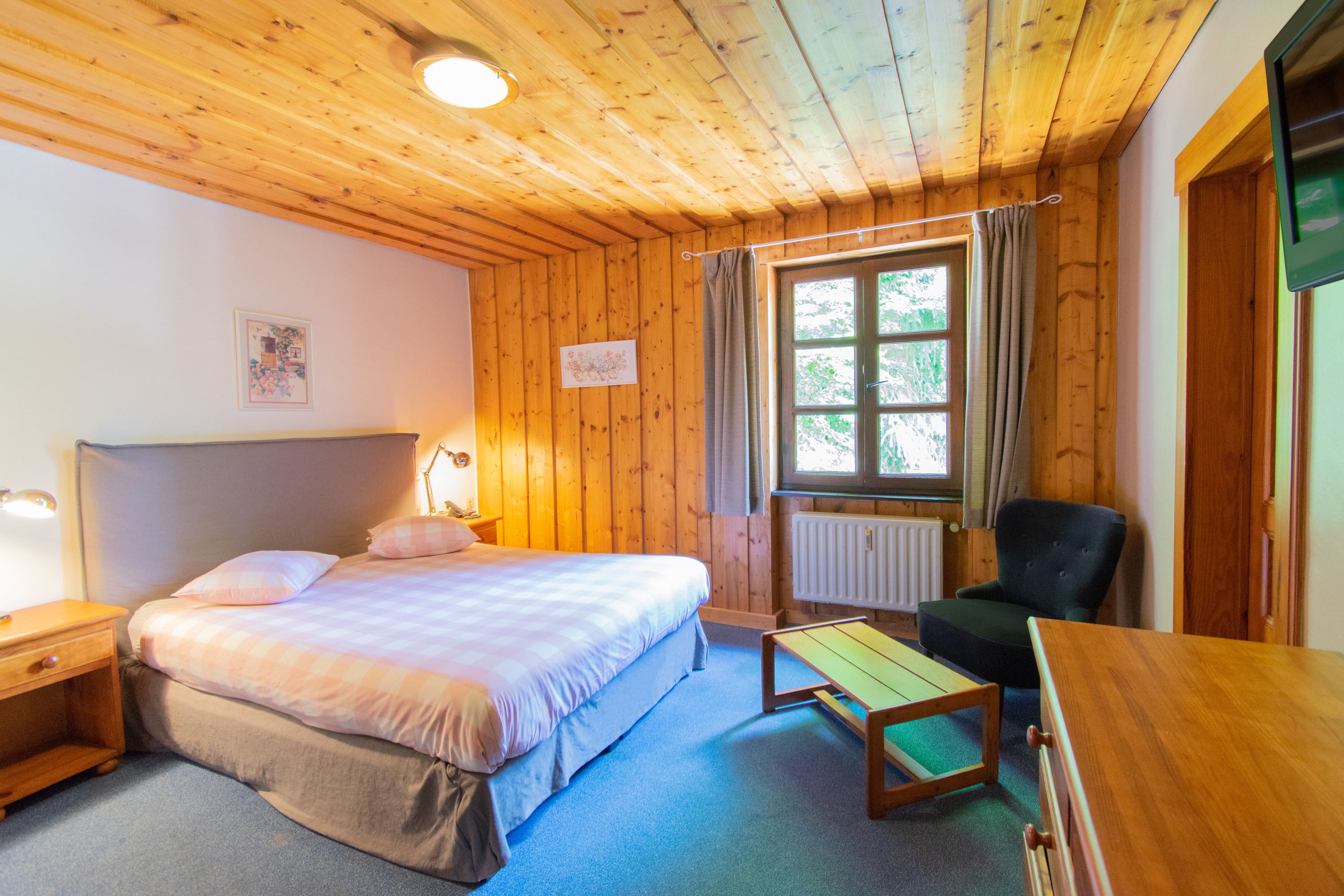 A wooden-paneled room with a bed, bedside table, chair, and a window with curtains.