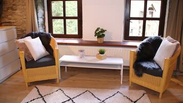 Une pièce chaleureuse avec deux fauteuils en osier, une table basse blanche, une petite plante en pot et une vue dégagée sur l’extérieur grâce à de grandes fenêtres.
