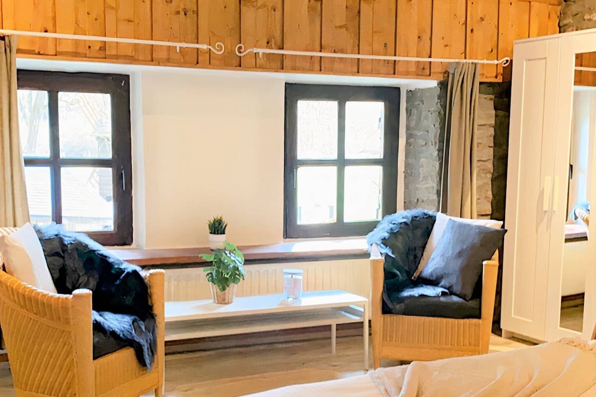 Ein gemütliches Zimmer mit Holzwänden und einem Bett mit weißer Decke. Zwei Korbstühle mit Kissen und ein kleiner Tisch mit einer Pflanze sind ebenfalls vorhanden.