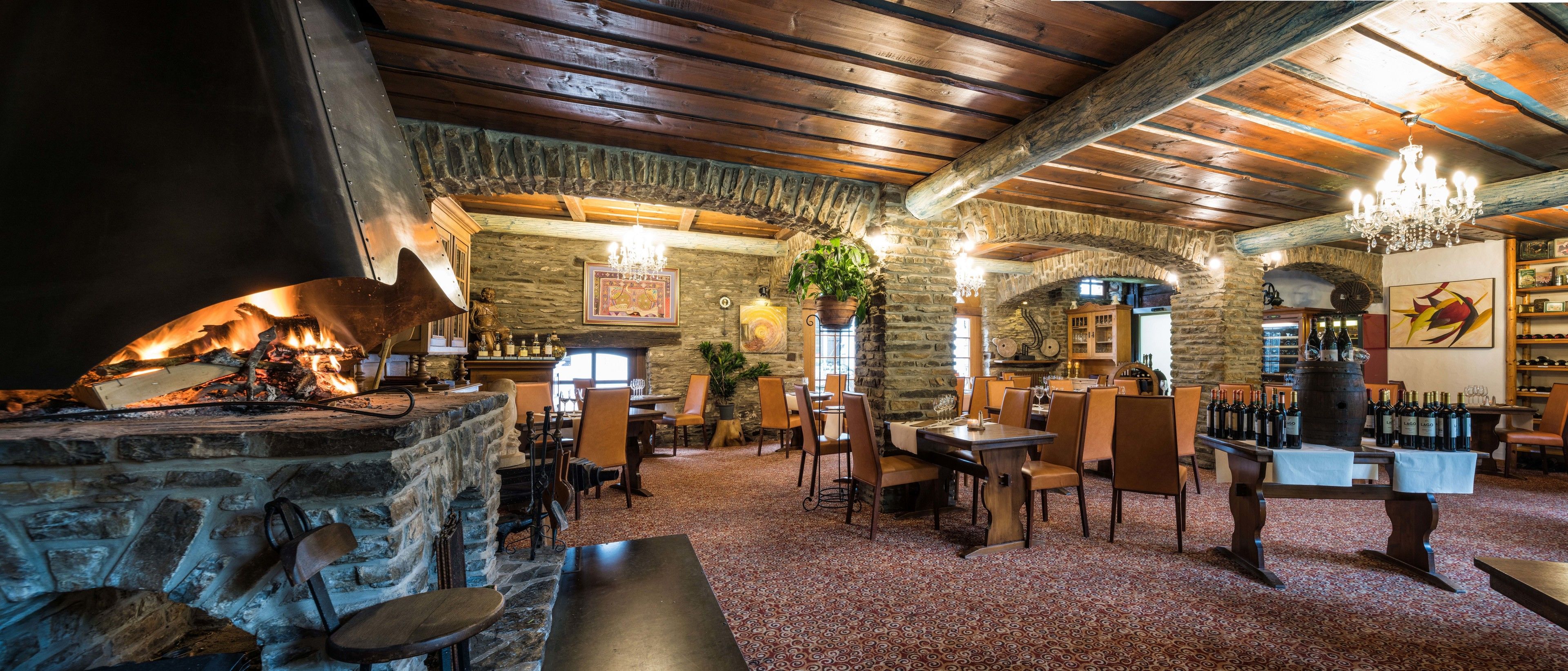 A cozy restaurant with stone walls, wooden beams, and a fireplace. Tables and chairs are arranged for dining.