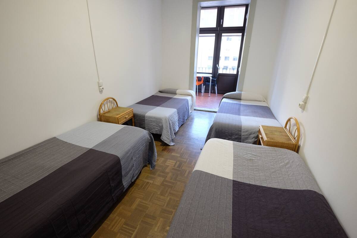 A room with three single beds, wooden headboards, and a small wooden table. A window with a view of the outside is visible.