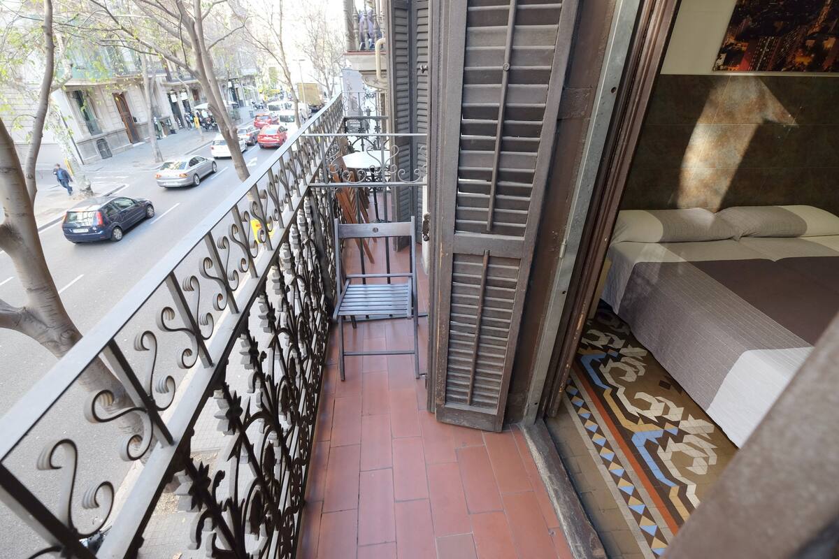 A narrow balcony with a metal railing, a small table, and a bed with a patterned bedspread.