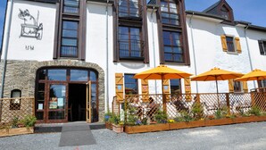 Ein Gebäude mit einer Stein-Fassade, großen Fenstern und Außenbestuhlung mit orangefarbenen Sonnenschirmen.