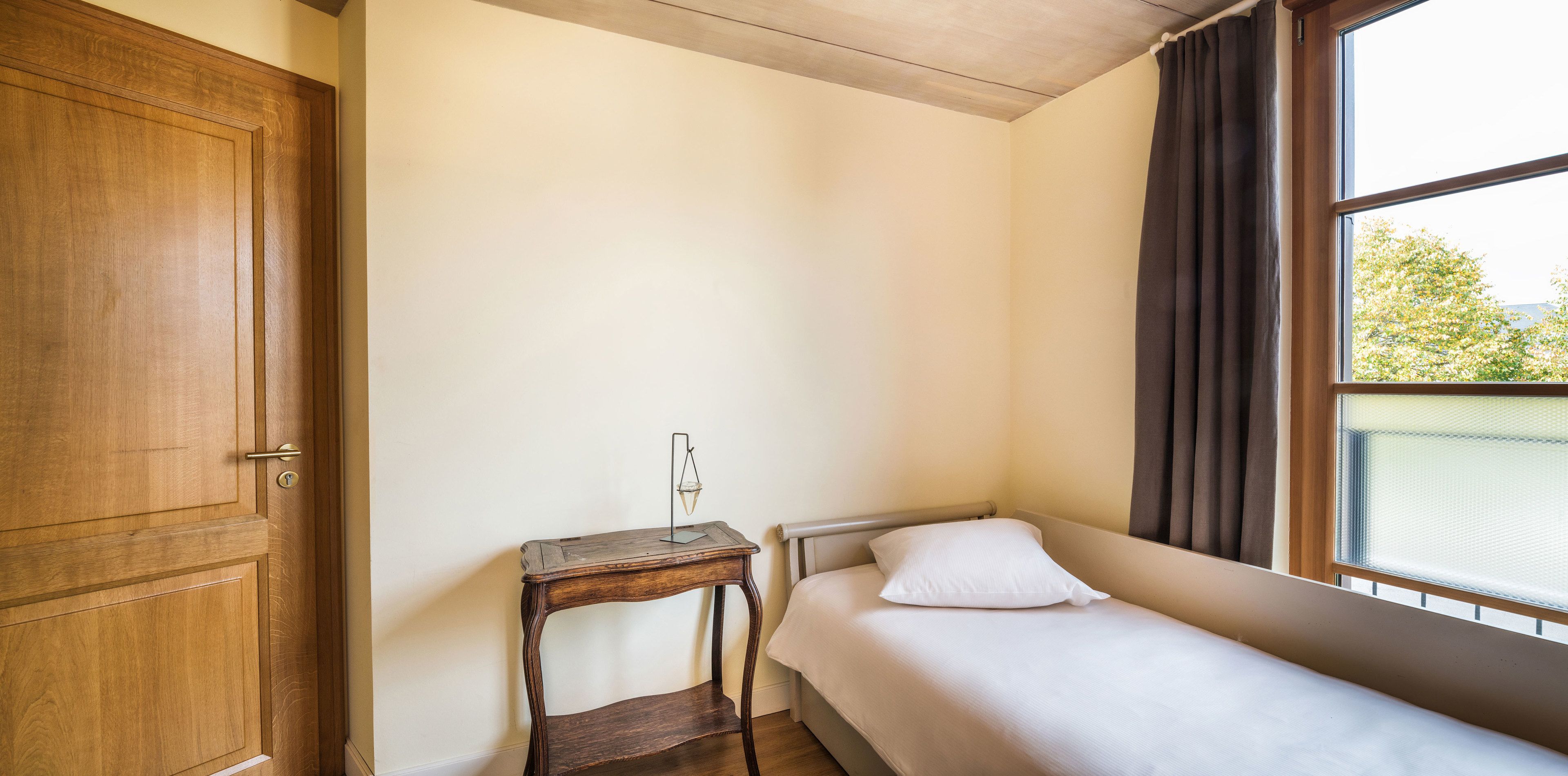 A small, single-bed room with a wooden door, a bedside table with a lamp, and a window with curtains.