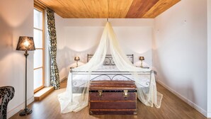 Ein Schlafzimmer mit Holzbalkendecke, einem Himmelbett, Nachttischlampen, einem Koffer und einer Stehlampe.