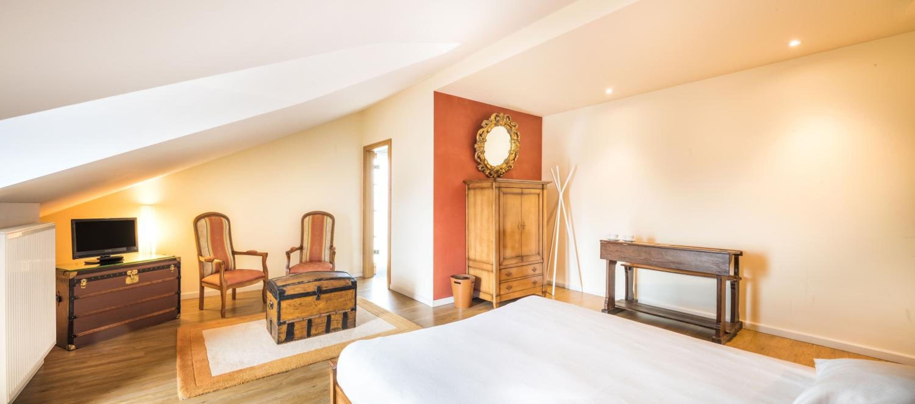 Een slaapkamer met een bed, een televisie, een houten kast, twee fauteuils en een spiegel.