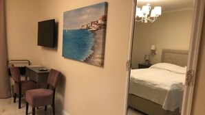 Et hotelværelse med en seng, et skrivebord med stol, et maleri af et kystlandskab og et spejl der reflekterer rummet.