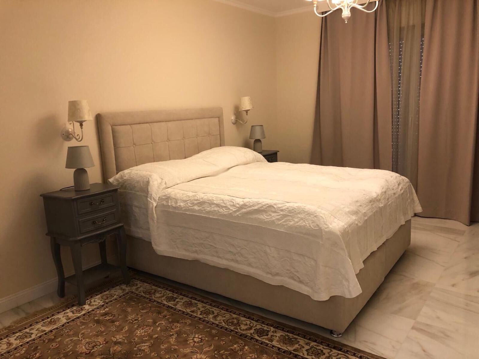 Een slaapkamer met een groot bed, twee nachtkastjes met lampen, een kamerkleed met patroon en een kroonluchter.