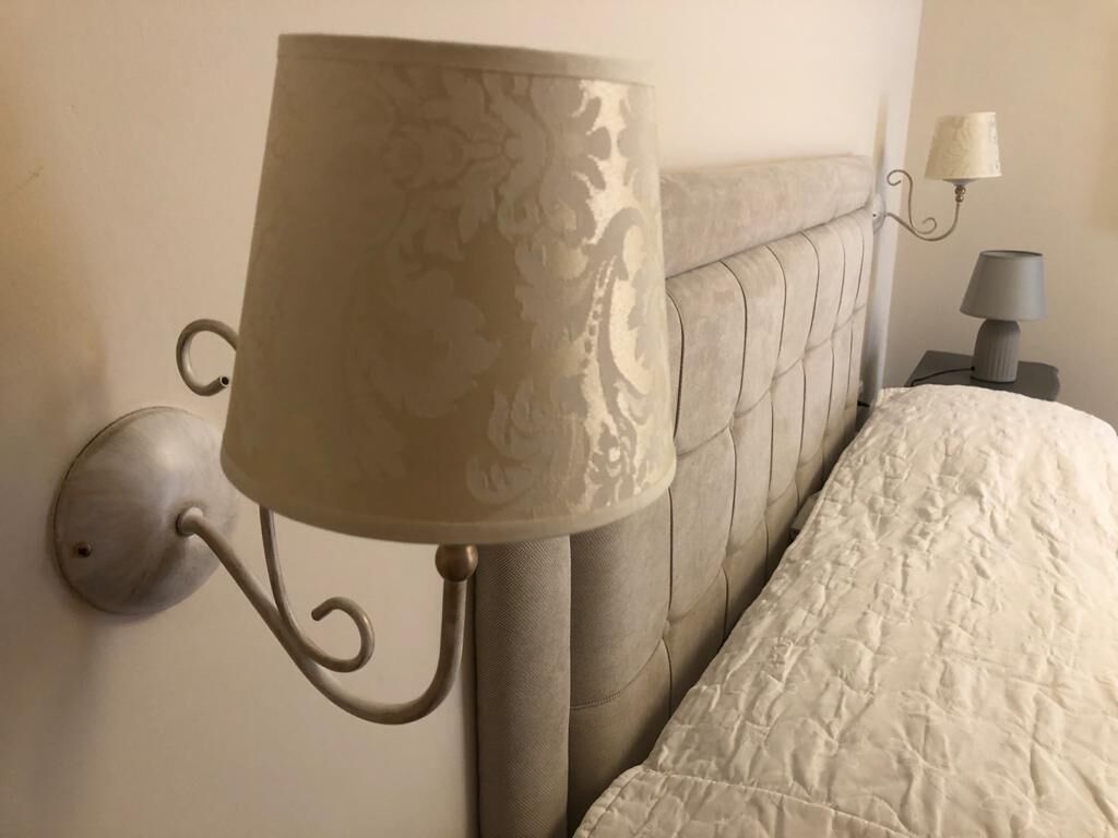 A wall-mounted lamp with a decorative shade, a bed with a quilted cover, and a bedside table with a lamp.
