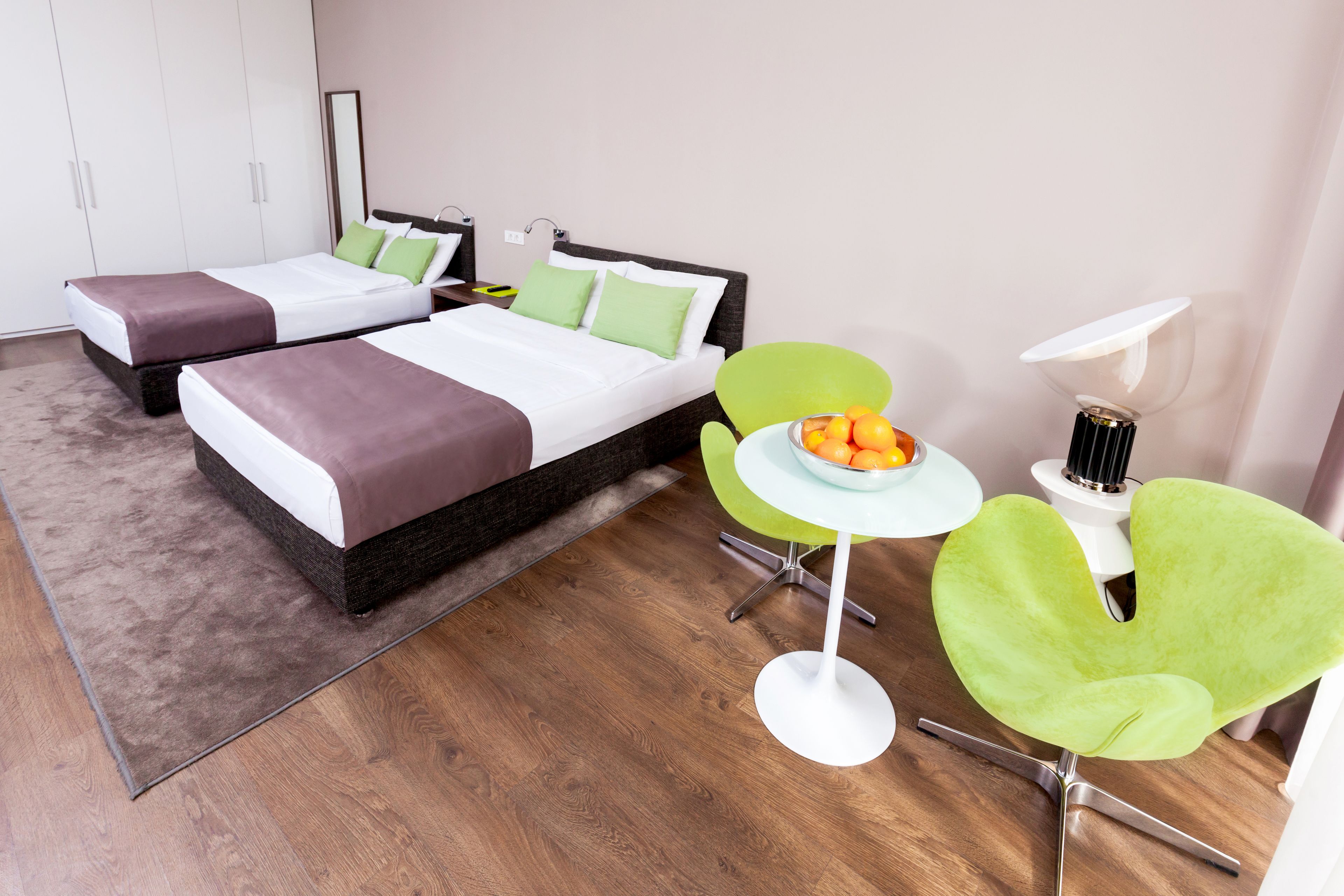 A modern hotel room with two beds, a small table with fruit, a green chair, and a lamp.
