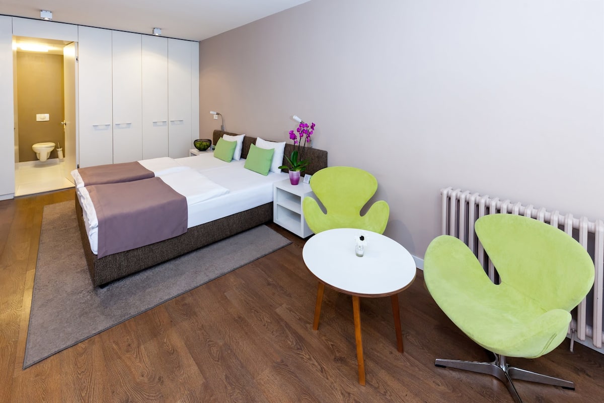Una habitación de hotel moderna con una cama grande, sillas verdes, una mesita pequeña y un radiador blanco.