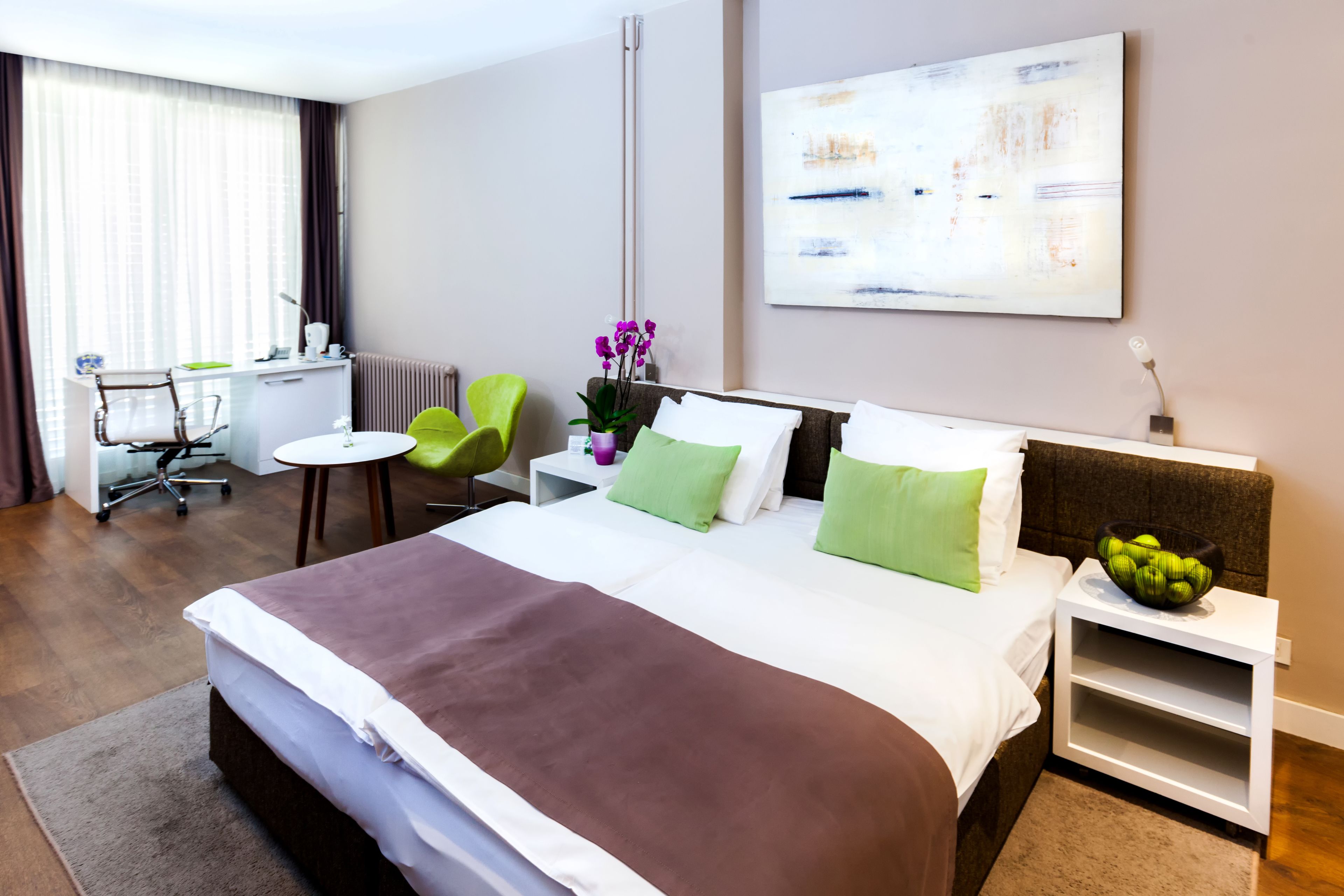 A hotel room with a large bed, a desk with a chair, a small table, and a wall-mounted shelf.