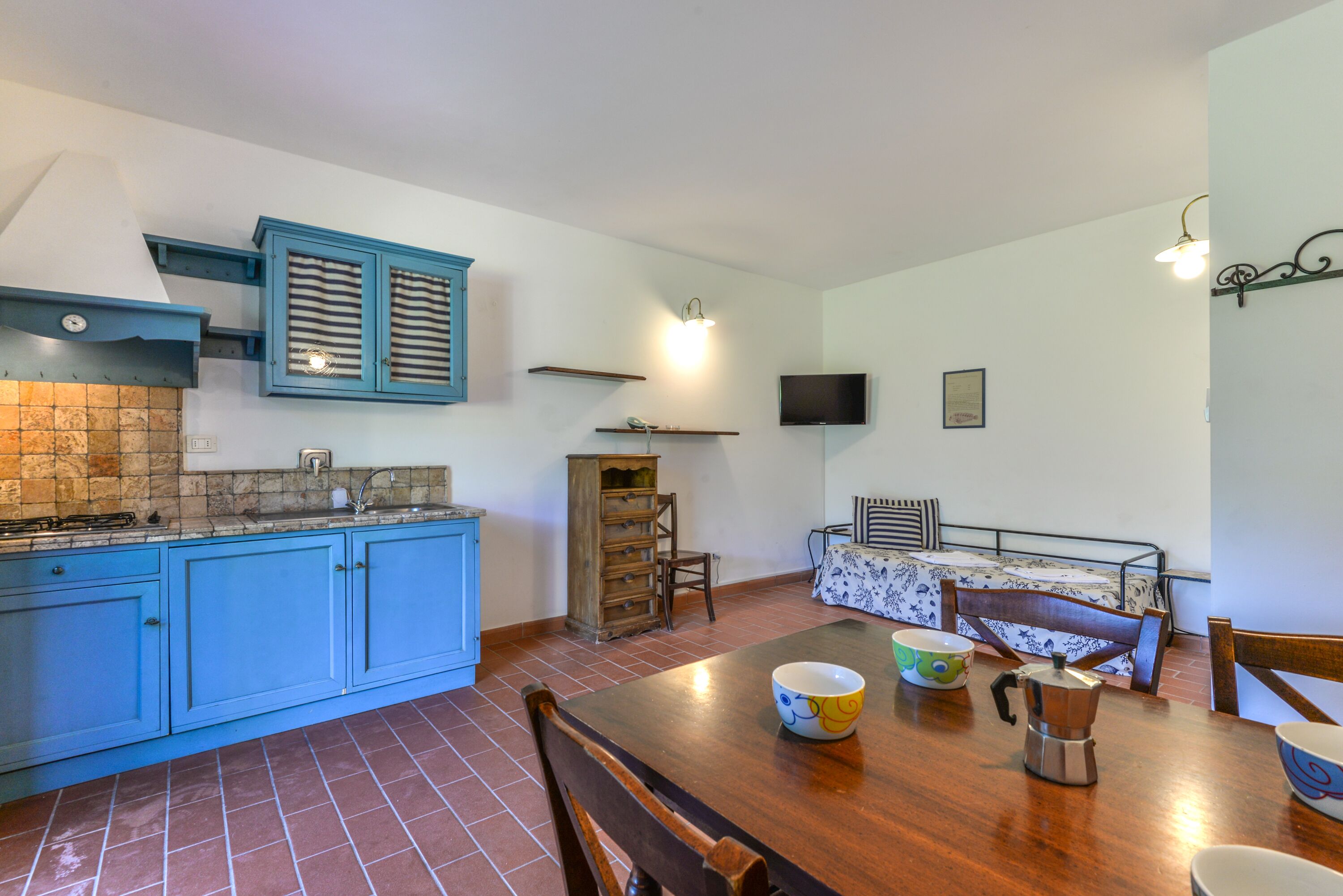 A compact kitchen with blue cabinets, a tiled backsplash, a dining table with chairs, and a small bed with a patterned bedspread.