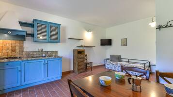 A compact kitchen with blue cabinets, a tiled backsplash, a dining table with chairs, and a small bed with a patterned bedspread.