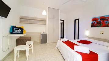 A hotel room with a bed, a desk, a chair, a small table, a TV, and a painting on the wall.