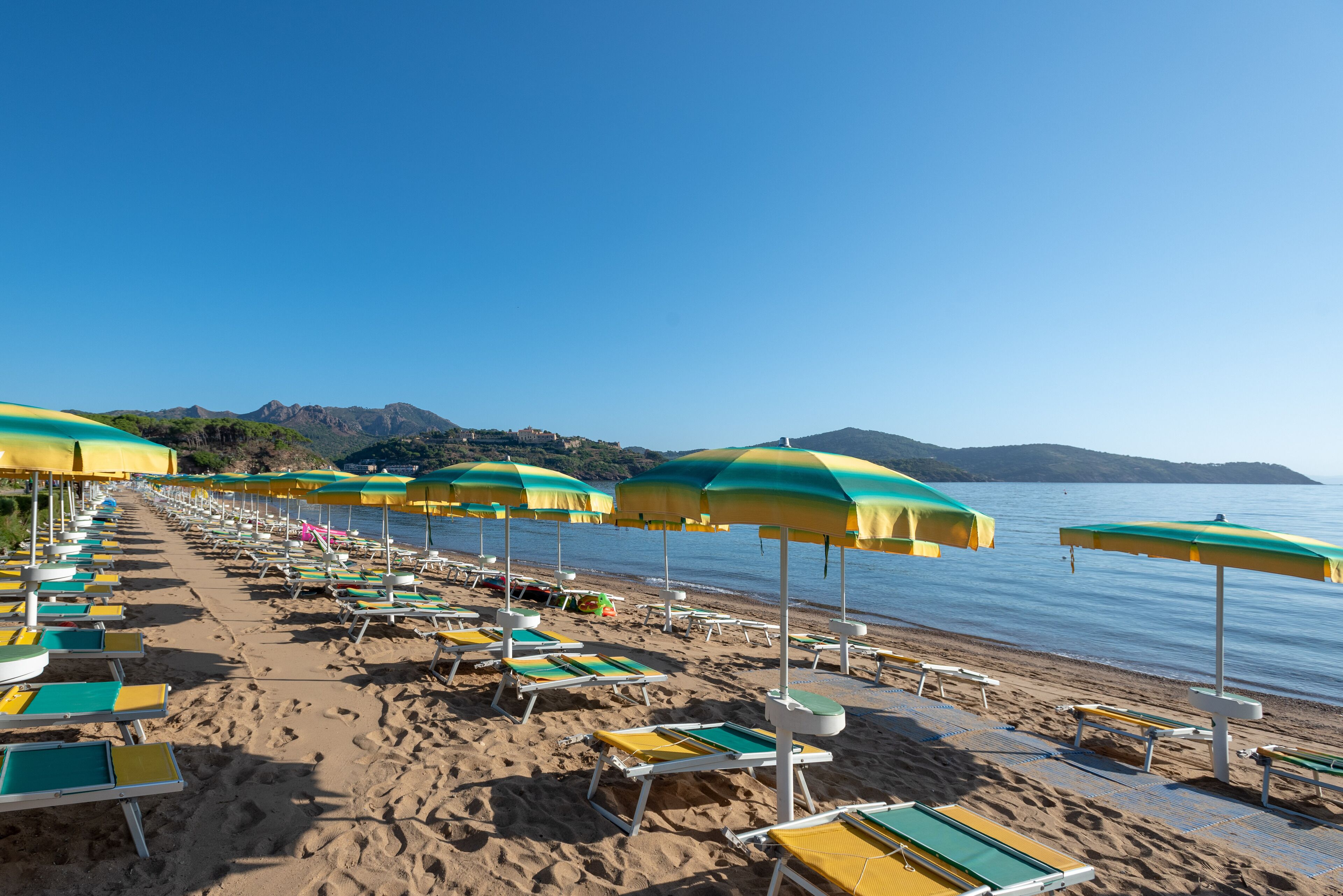 A beach with rows of sun loungers and umbrellas, a clear sky, and mountains in the background.