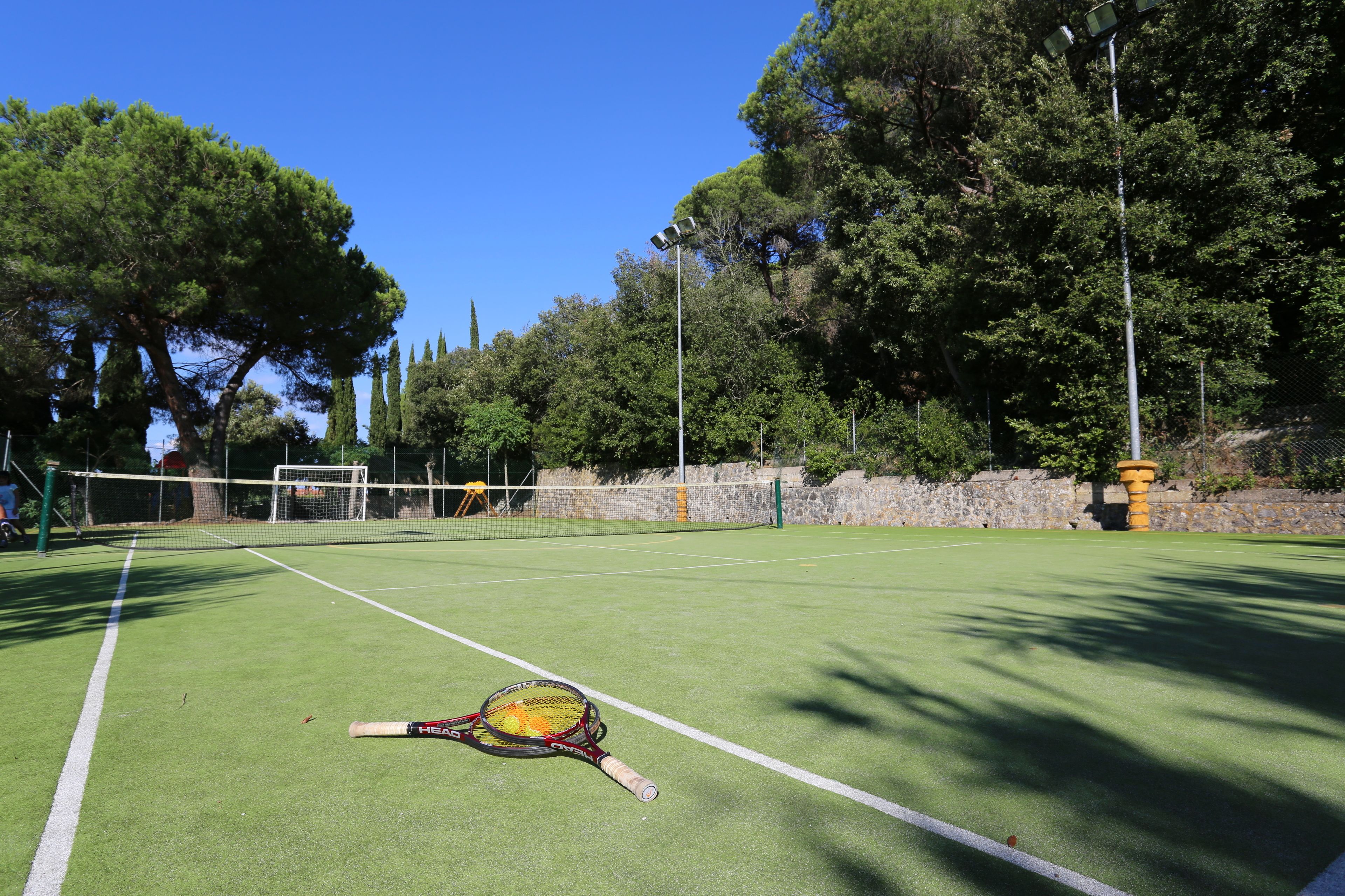Una cancha de tenis con red, rodeada de árboles y un muro de piedra.