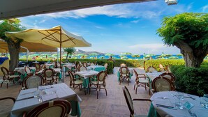 Un área de comedor al aire libre con mesas y sillas, con vista a una playa con sombrillas y gente.