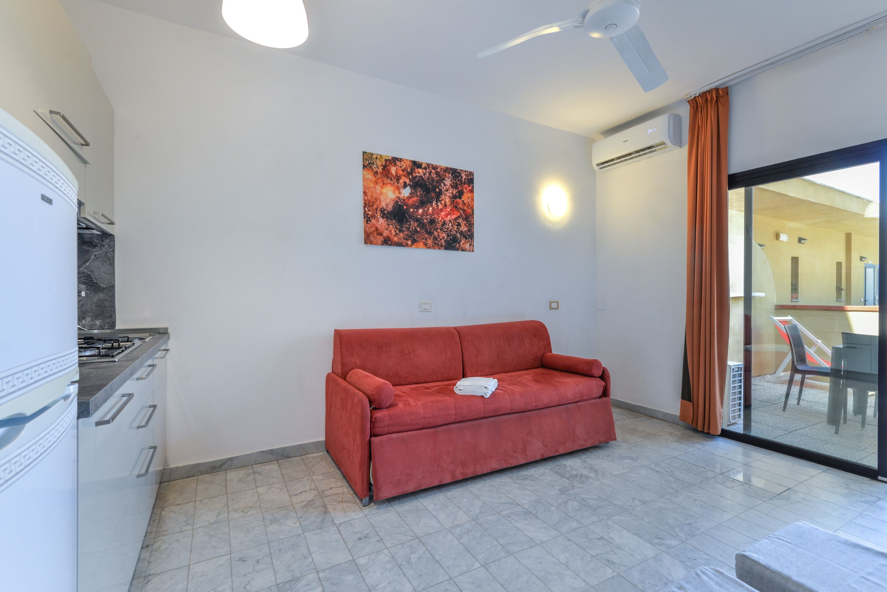 Ein modernes Wohnzimmer mit einem roten Sofa, einer Küchenzeile inklusive Gaskochfeld und einer Glastür zum Außenbereich.
