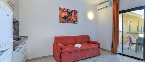 A modern living room with a red sofa, a kitchen area with a gas stove, and a glass door leading to an outdoor area.