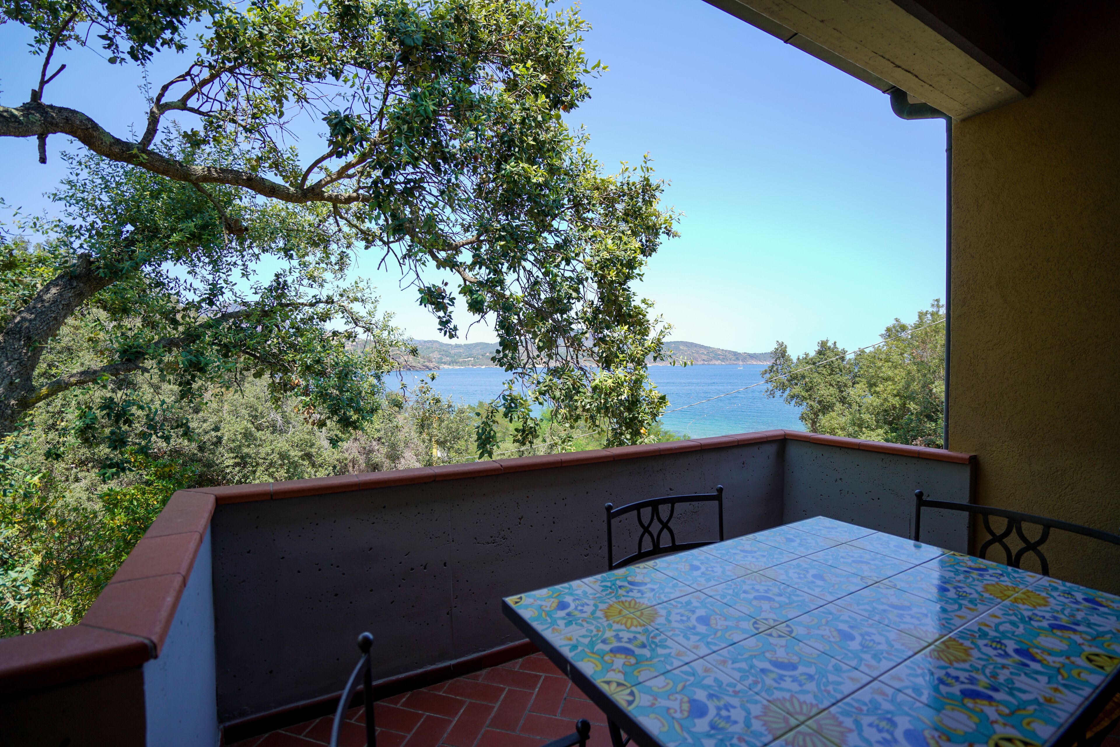 A balcony with a tiled table and chairs, overlooking a body of water and greenery.