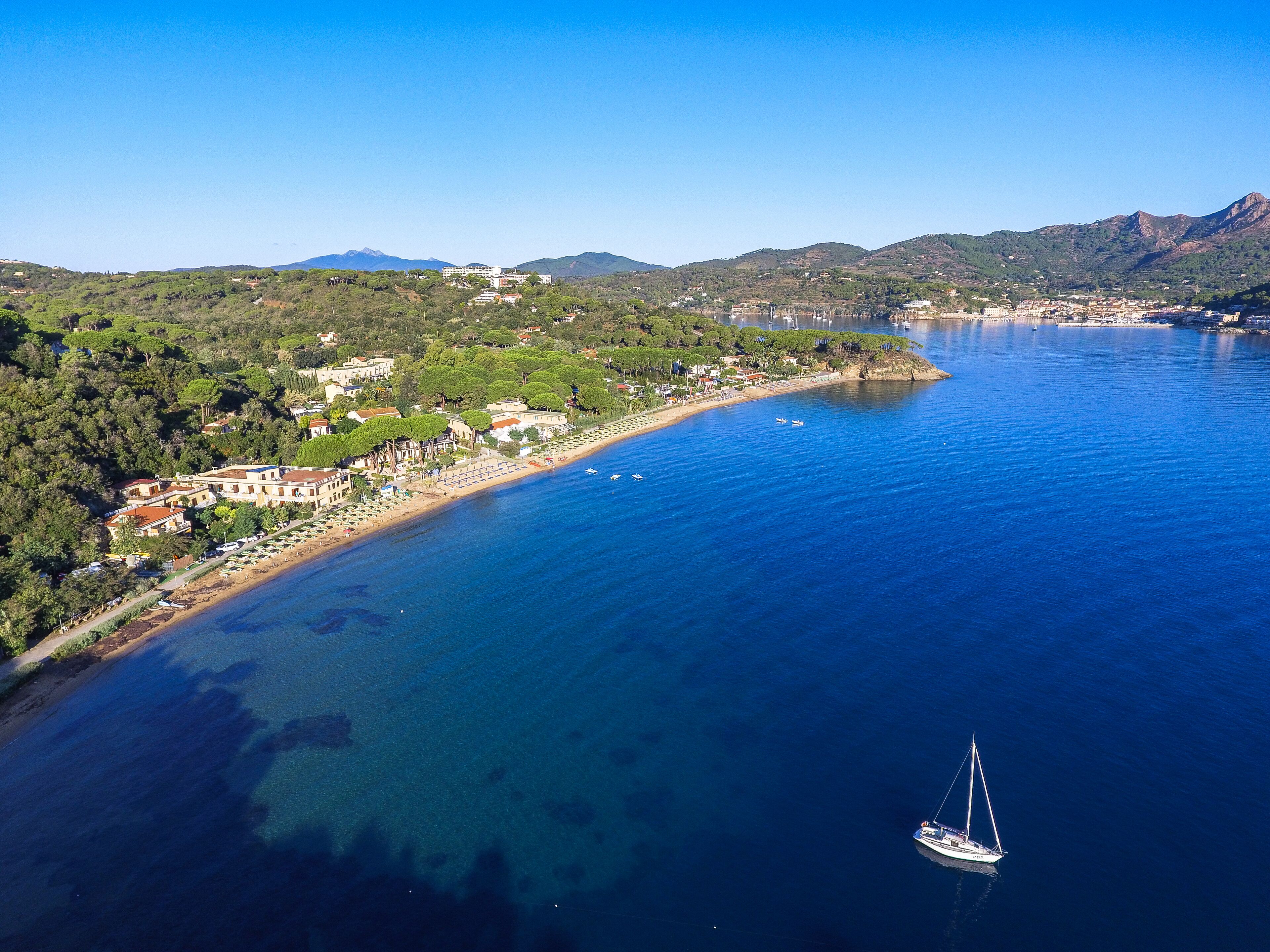 An aerial view of a coastal town with a marina, clear blue waters, and a sailboat.
