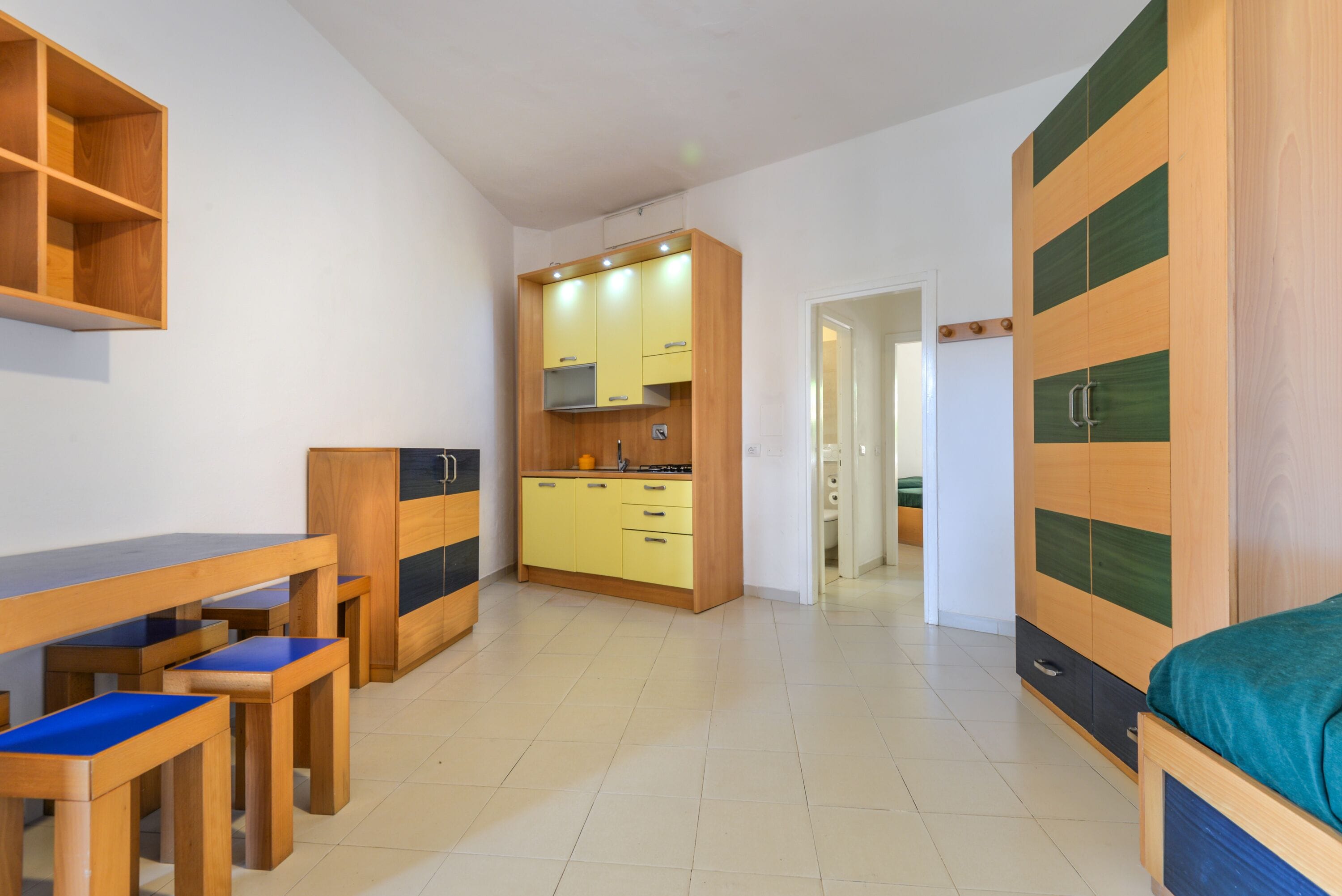 A compact room with a kitchenette, a bed, a wooden desk with chairs, and a wardrobe.