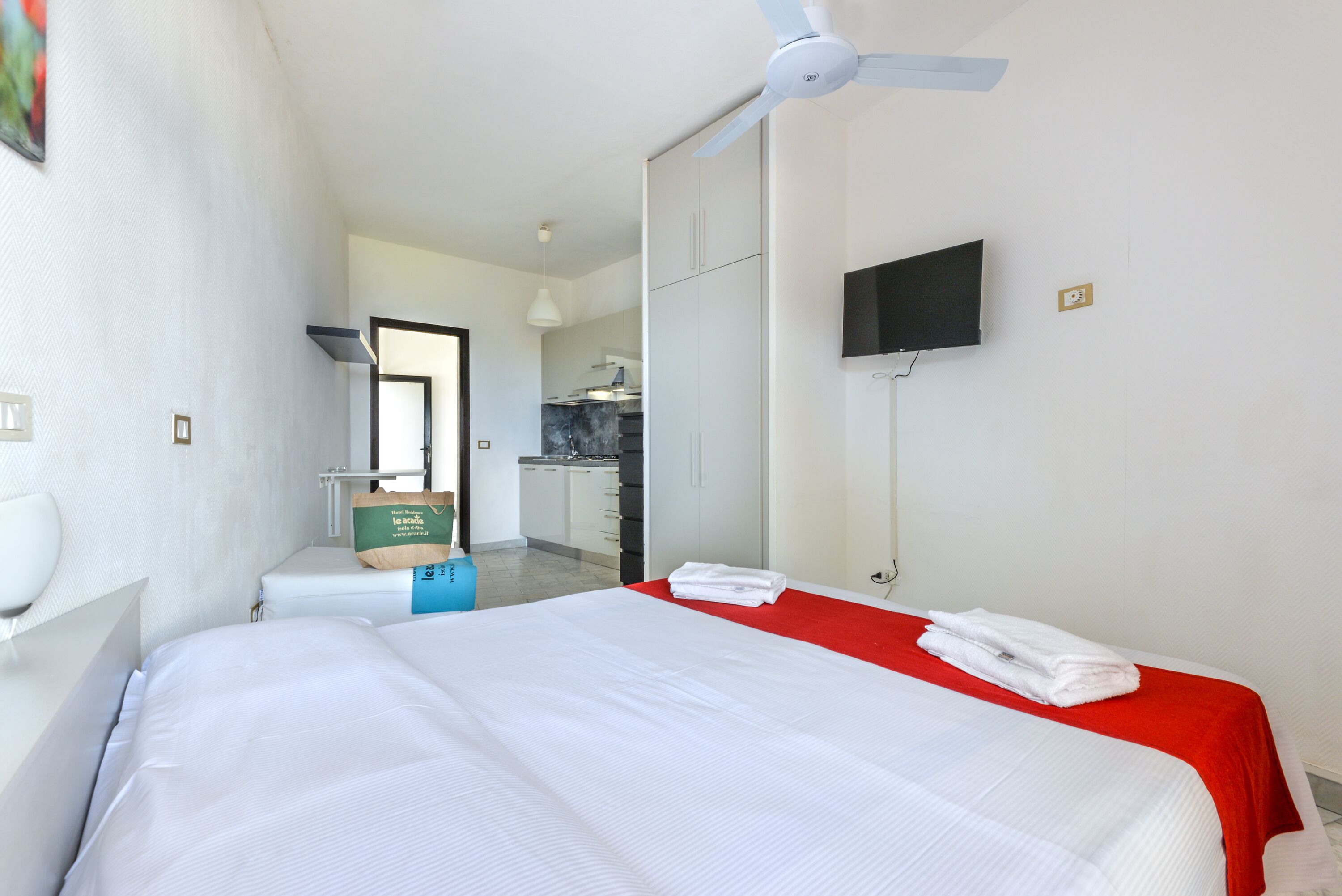 Una habitació d'hotel amb un llit, una paret vermella decorativa, un ventilador de sostre, una televisió i vista a la zona de la cuina.