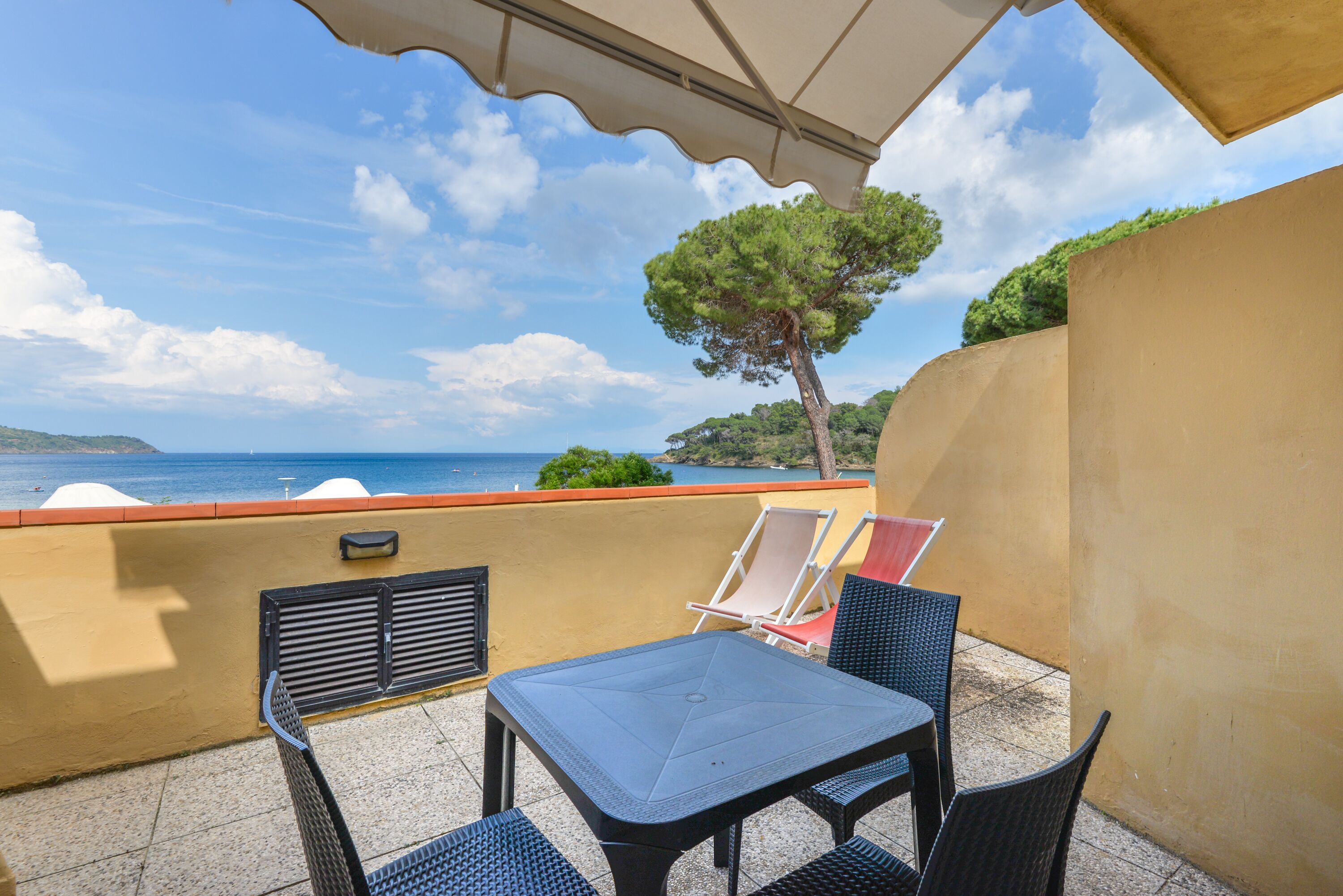 A balcony with a table and chairs, overlooking a scenic view of the sea and a tree.