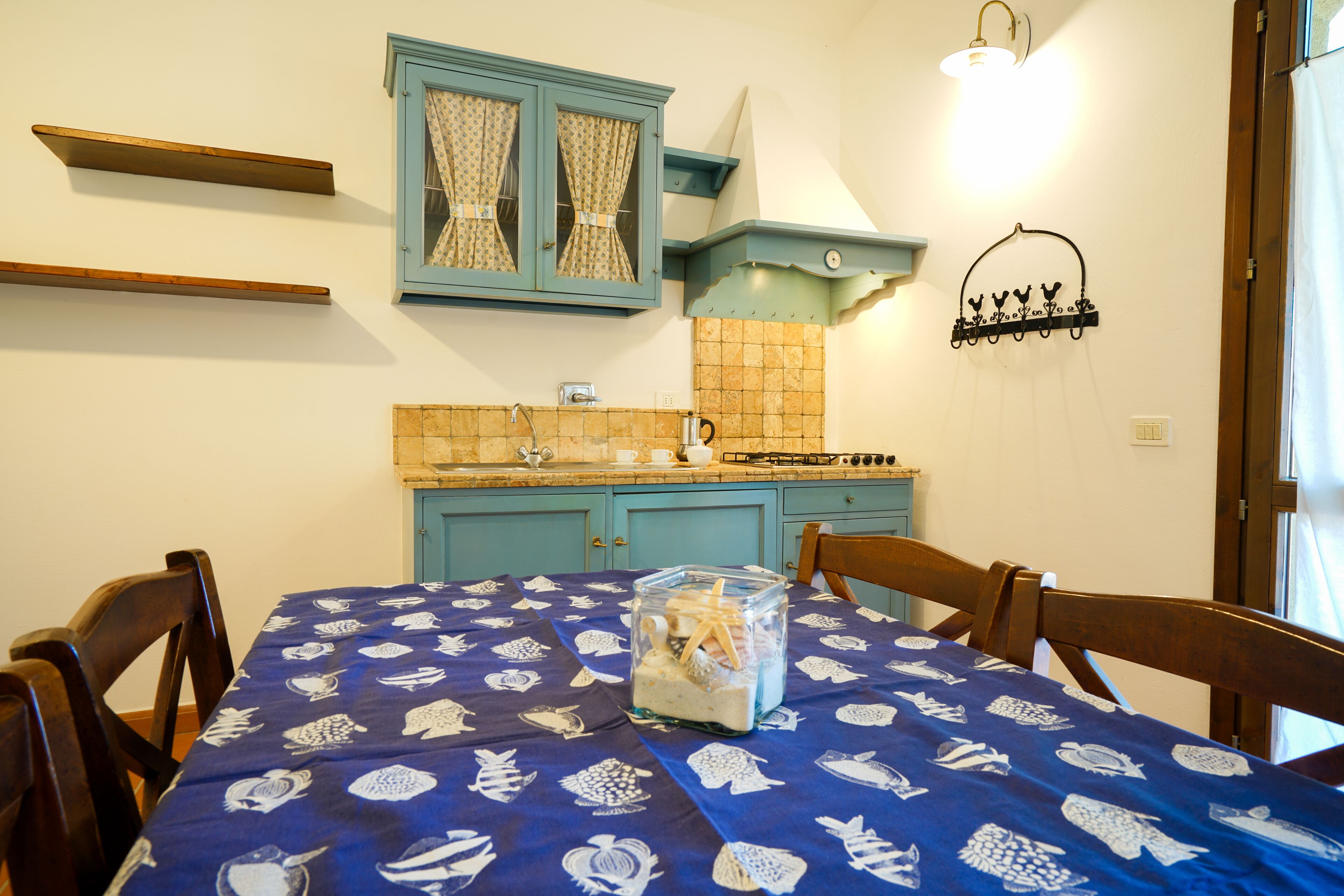 A dining area with a table covered in a blue tablecloth with white patterns, wooden chairs, a blue cabinet with curtains, a tiled backsplash, and a hanging light fixture.