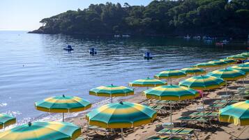 Uma praia com fileiras de guarda-sóis e espreguiçadeiras, um mar calmo e barcos ao fundo.