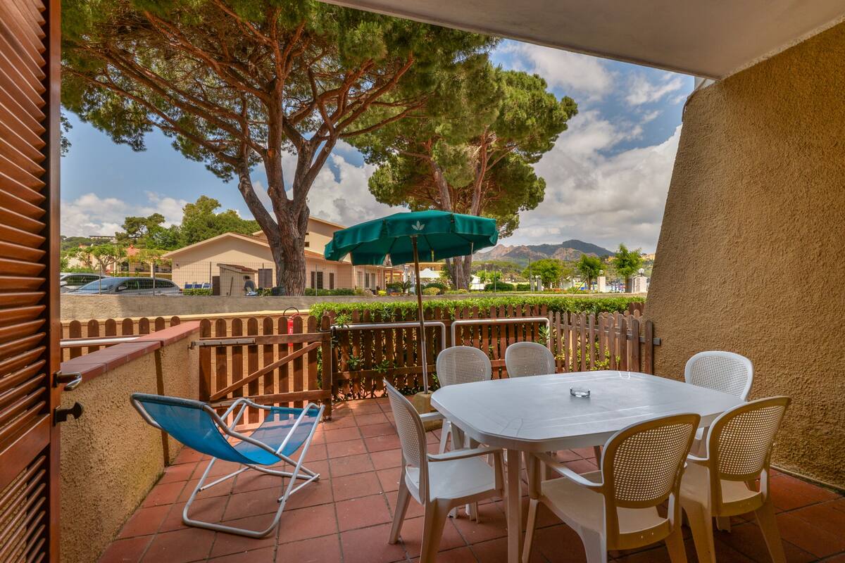 En udendørs terrasse med et bord og stole, en blå klapstol, en grøn parasol og udsigt til træer og bjerge.