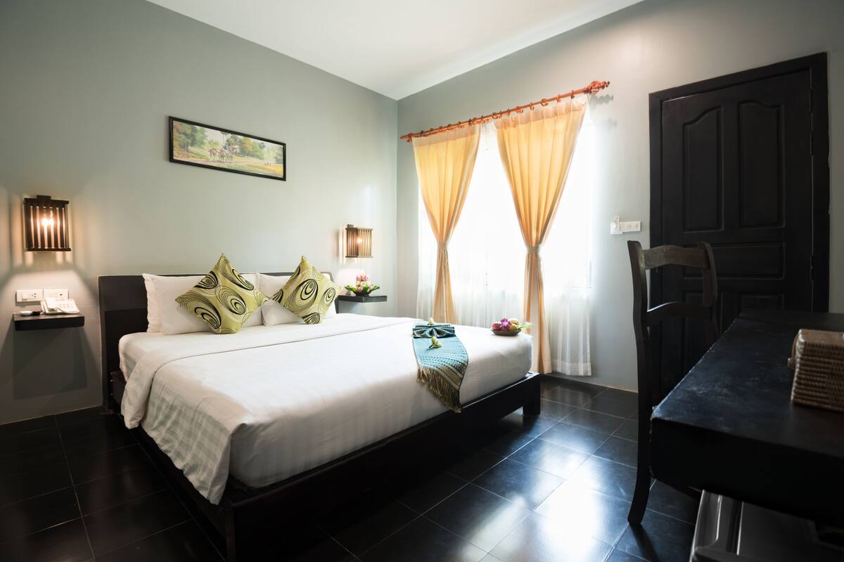 A bedroom with a bed, bedside lamps, a window with curtains, a dining table, and a chair.