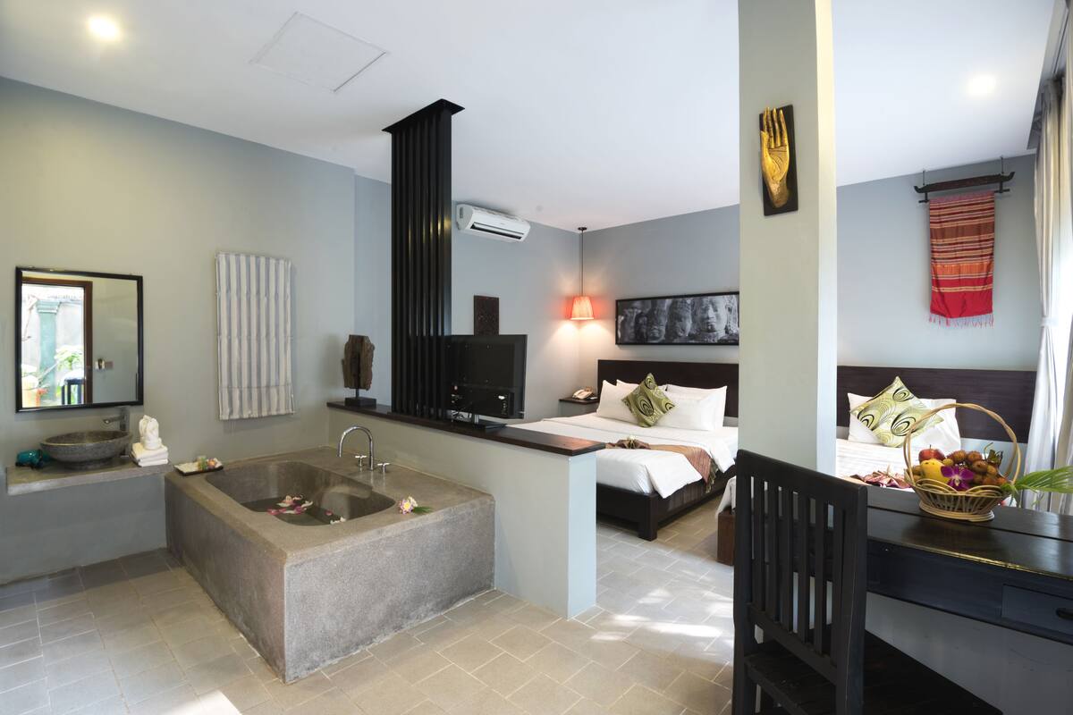 A modern bathroom with a large bathtub, a sink, and a mirror. There is a bed with pillows and a bedside table with a lamp.