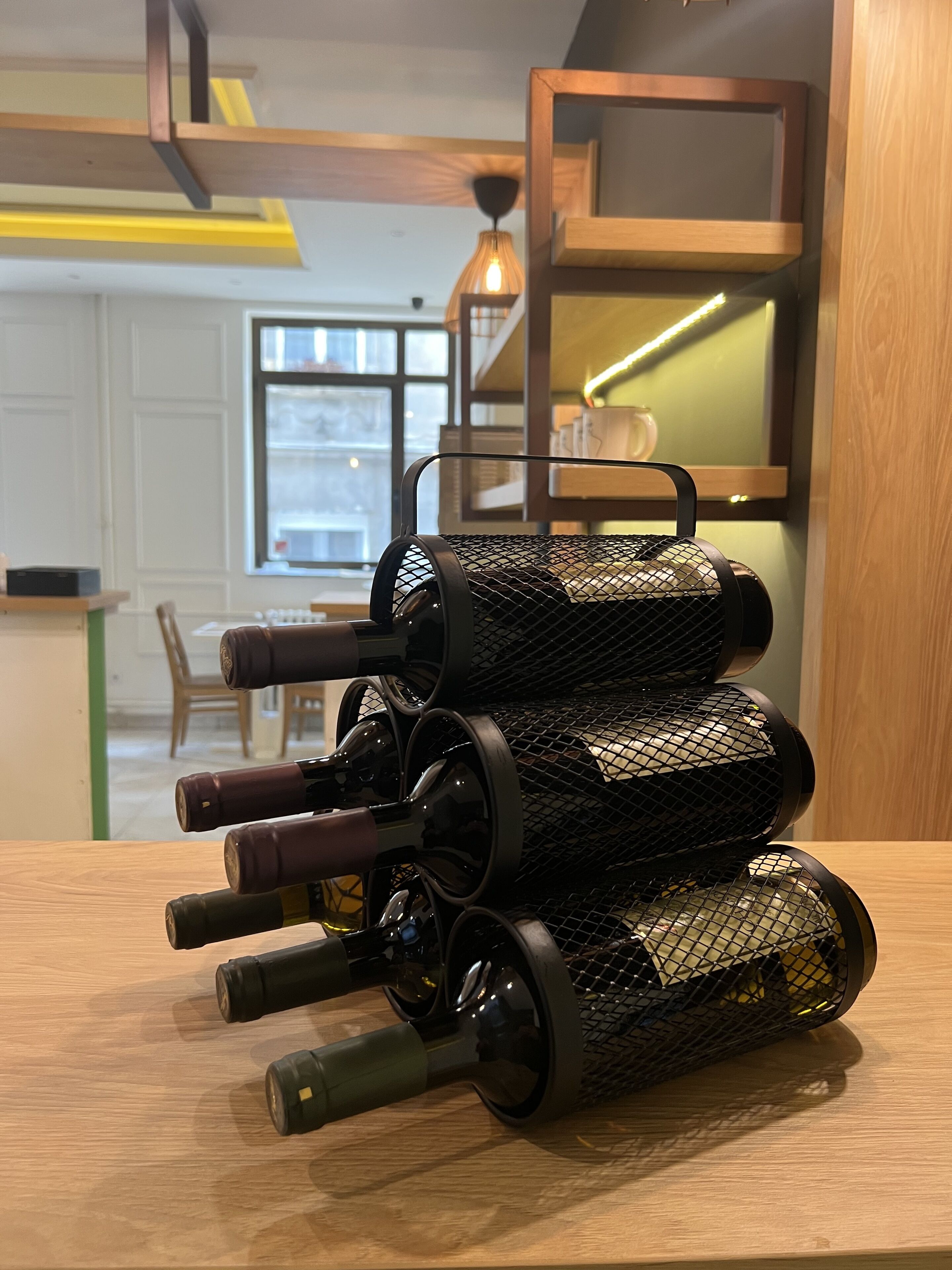 A wine rack with multiple bottles on a wooden table in a room with a modern design, including a hanging light fixture and a window with a view.