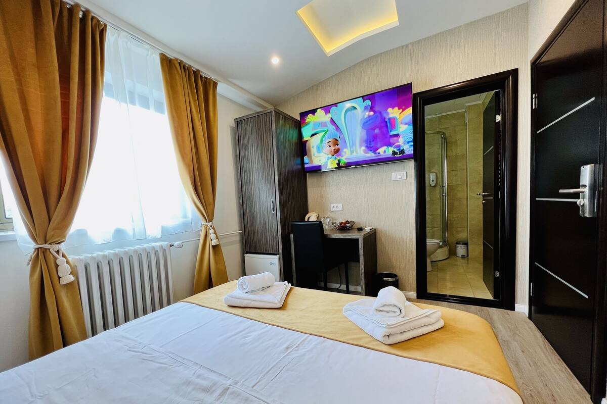 Quarto de hotel com cama, escrivaninha, televisão e banheiro visível por uma porta aberta.