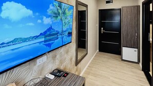 Una habitación de hotel moderna con un televisor de pantalla plana grande montado en una pared con textura, un escritorio de madera con un teléfono y controles remotos, y una puerta negra elegante.