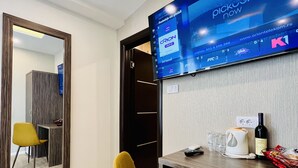 Una habitación de hotel moderna con un televisor de pantalla plana grande montado en la pared, mostrando una interfaz digital. Hay un escritorio de madera con una tetera blanca, una botella de vino y algunos aperitivos. Una silla color mostaza está colocada frente al escritorio. Un espejo grande se encuentra en la pared opuesta al escritorio.