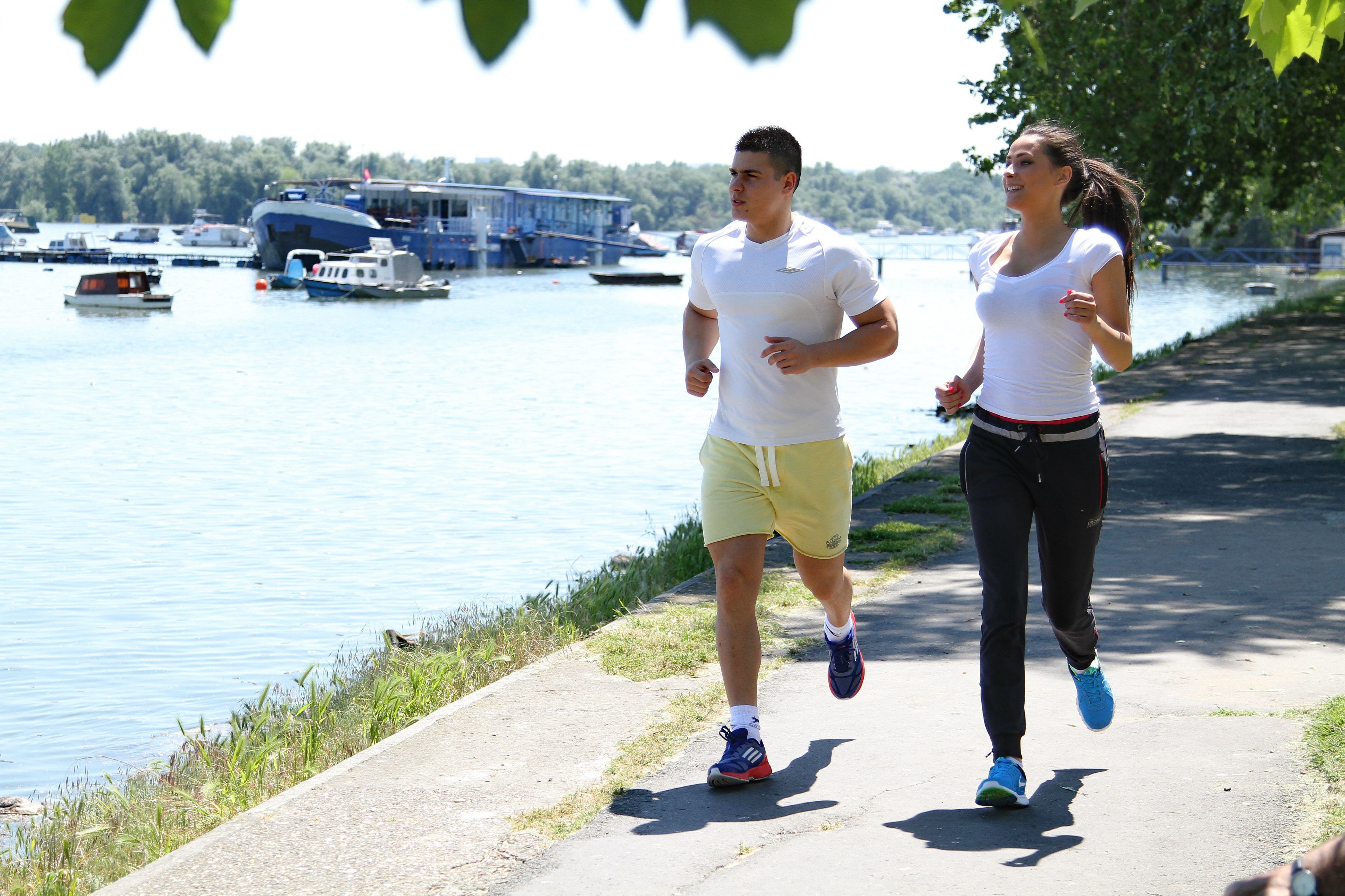 Dos personas trotando por un sendero junto a un cuerpo de agua, con barcos y un muelle al fondo.