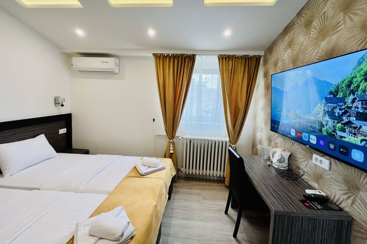 Quarto de hotel com cama, secretária, televisão e janela com cortinas.