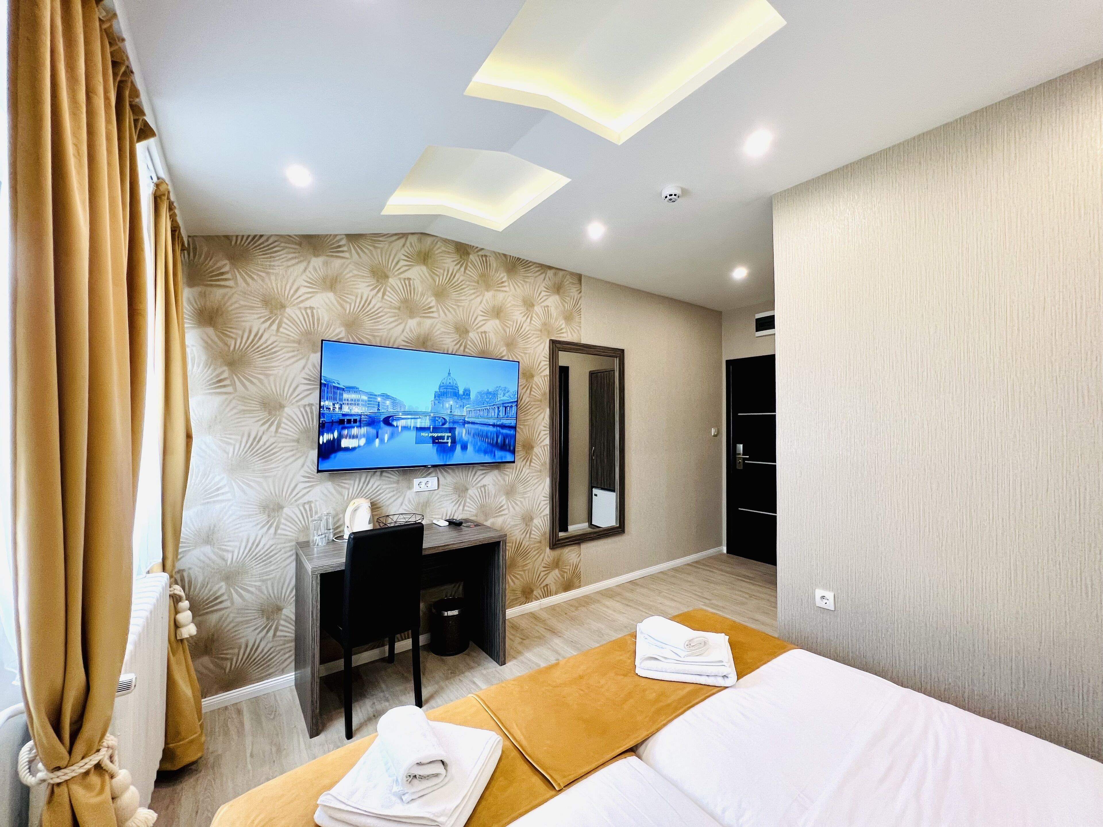 A hotel room with a flat-screen TV mounted on the wall, a desk with a chair, a bed with a yellow bed runner, and a large mirror.